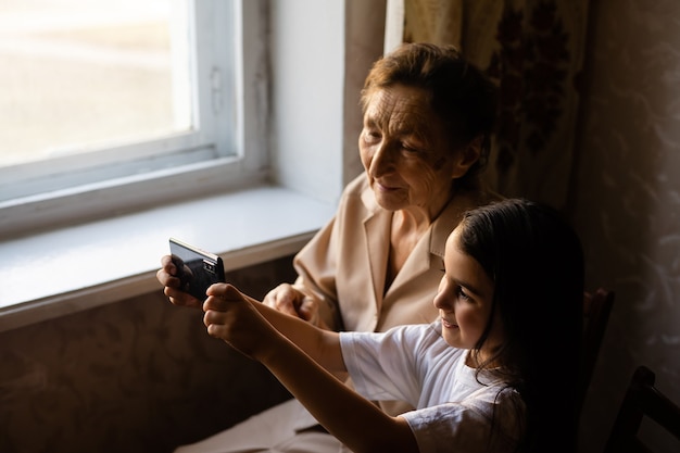 Prababka siedzi z prawnuczką i zagląda do smartfona. Babcia i dziecko robią sobie selfie na smartfonie. Babcia z wnuczką zaglądają do telefonu.