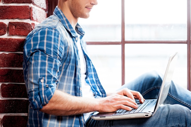 Praca na laptopie. Widok z boku młodego mężczyzny pracującego na laptopie siedząc na parapecie