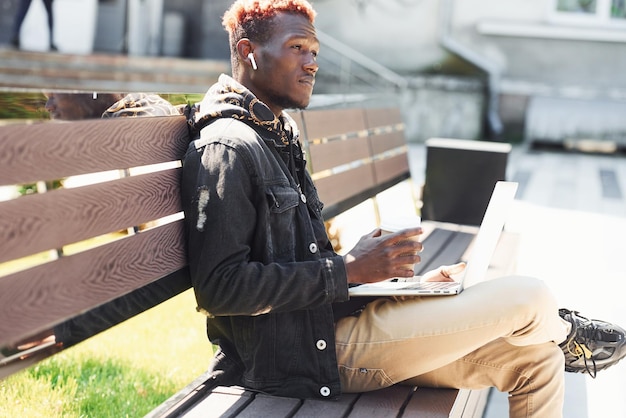 Praca przy użyciu laptopa Młody Afroamerykanin w czarnej kurtce na zewnątrz w mieście, siedząc na ławce