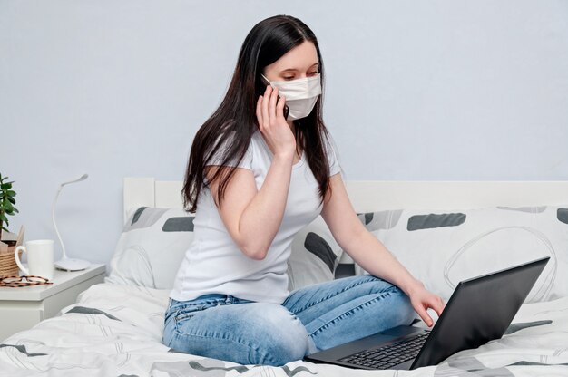Praca w domu lub na odległość Nauka online na kwarantannie. Kobieta w masce ochronnej zdalnie pracuje na łóżku za pomocą laptopa i rozmawia przez telefon.