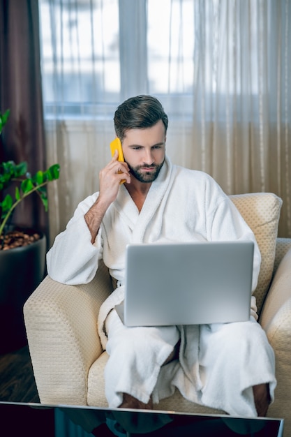 Praca W Domu. Młody Brodaty Mężczyzna W Białym Szlafroku Pracuje Na Laptopie I Rozmawia Przez Telefon