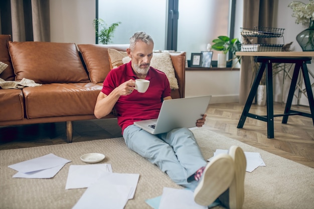 Praca w domu. Siwy mężczyzna siedzi na podłodze i pracuje na laptopie