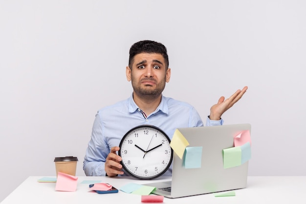 Praca w godzinach nadliczbowych Nieszczęśliwy zmęczony człowiek pracownik siedzący w biurze w miejscu pracy trzymający zegar podnoszący rękę w oburzonym geście przesłuchania karteczki dookoła przypominające o terminie strzał studio na białym tle