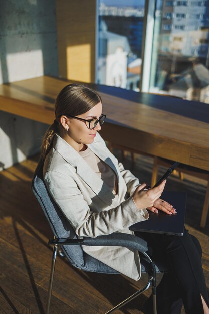 Praca w nowoczesnym biurze Pozytywna młoda bizneswoman w zwykłych ubraniach i okularach siedzi w biurze z telefonem komórkowym podczas pracy nad zdalnym projektem przy użyciu laptopa