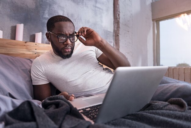 Praca w sypialni. Afrykański młody człowiek skoncentrowany w okularach i za pomocą laptopa, leżąc w łóżku.