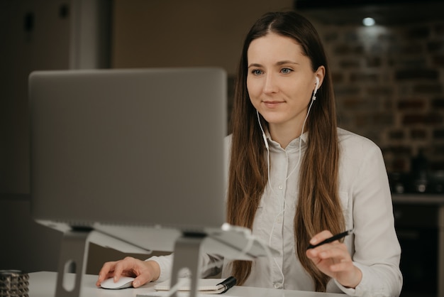 Praca zdalna. Kaukaska brunetki kobieta z hełmofonami pracuje zdalnie na jej laptopie. Bizneswoman w białej koszula robi biznesowi przy jej domowym miejscem pracy.