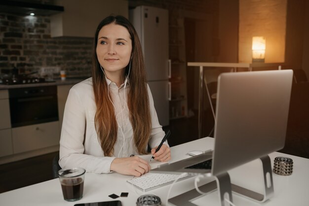 Praca zdalna. Kaukaska kobieta ze słuchawkami pracującymi zdalnie na swoim laptopie. Szczęśliwa brunetka z uśmiechem robiąc notatki podczas odprawy biznesowej online w swoim przytulnym domowym miejscu pracy.