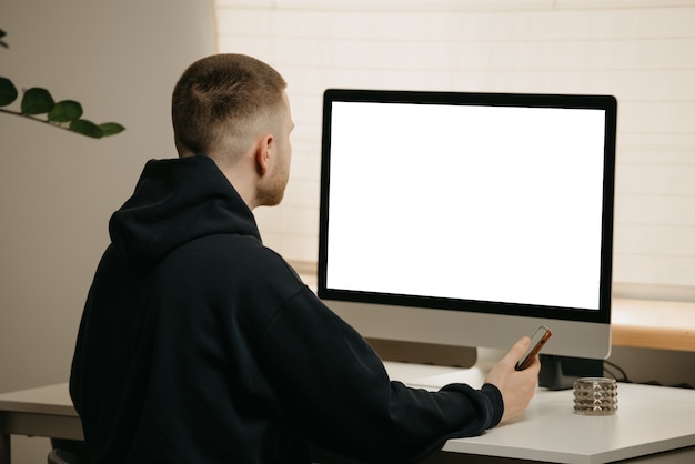 Praca zdalna. Widok biznesmena z tyłu działa zdalnie przy użyciu komputera typu „wszystko w jednym”. Facet pracuje, trzymając smartfon z domu.