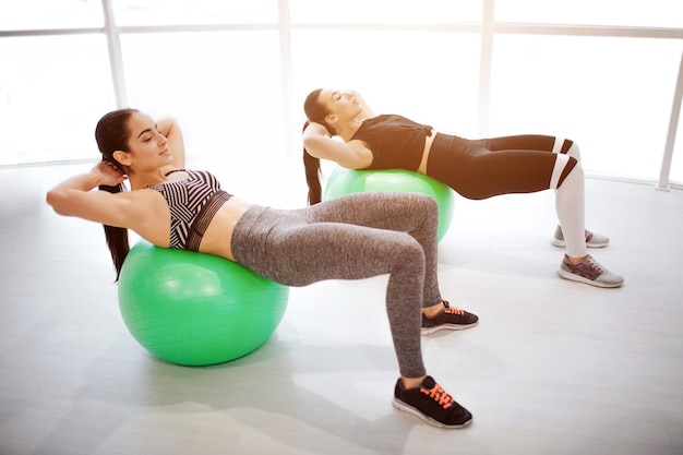 Pracowite młode kobiety leżące na zielonych piłkach fitball i ćwiczą abd. Trzymają ręce za głową. Modelki w oknie