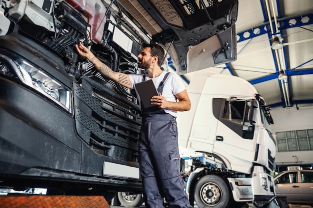 Pracowity, wytatuowany, brodaty mechanik, oparty na ciężarówce i sprawdzający silnik, stojąc w garażu firmy importowo-eksportowej.