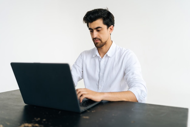 Pracowniany portret poważnego skupionego biznesmena w koszuli pracującego na laptopie wpisując na klawiaturze siedzącego przy biurku na białym, odizolowanym tle