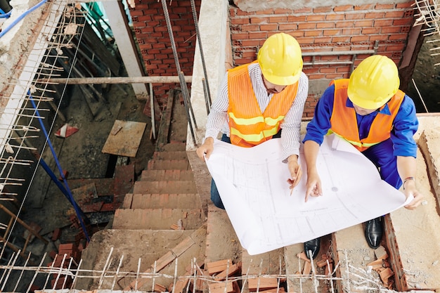 Zdjęcie pracownicy budowlani siedzący na schodach wewnątrz budynku i omawiający szczegóły projektu, widok z góry