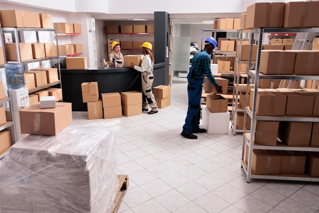 Pracownicy działu wysyłek wybierają paczki do dostawy i umieszczają karton na biurku. Pracownicy magazynu w kombinezonach ochronnych i hełmach przygotowujący ładunek do wysyłki