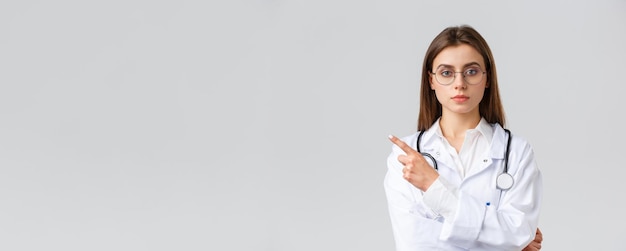 Pracownicy opieki zdrowotnej medycyna ubezpieczenie i koncepcja pandemii covid19 Poważnie wyglądająca profesjonalna lekarka pielęgniarka w białych fartuchach i okularach wskazująca palcem w lewo na baner lub informacje o klinice