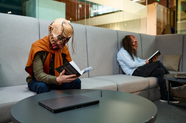 Pracownicy zombie czytają książki podczas przerwy w pracy, siedząc na kanapie w holu budynku biznesowego. Biuro typu open space