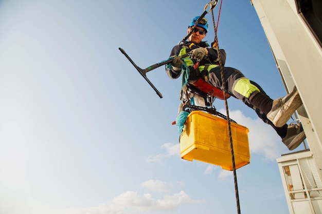 Pracownik Alpinizmu Przemysłowego Wisi Nad Budynkiem Mieszkalnym Podczas Mycia Przeszklenia Elewacji Zewnętrznej. Robotnik Z Dostępem Linowym Wisi Na ścianie Domu