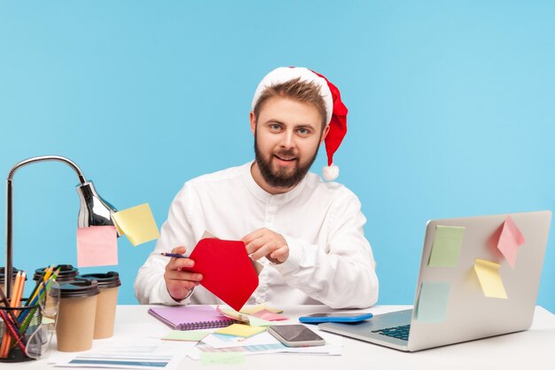 Pracownik biurowy przystojny brodaty mężczyzna w kapeluszu świętego mikołaja siedzi w miejscu pracy i podpisuje pocztówki z gratulacjami z ferii zimowych robi niespodziankę Kryty studio strzał na białym tle na niebieskim tle