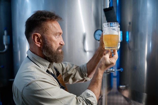 Pracownik browaru patrzący na świeżo zrobione piwo w szklanym kubku. Koncepcja procesu technologicznego browaru
