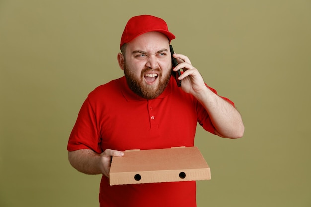 Pracownik Dostawy W Czerwonej Czapce Pusta Koszulka Mundurze Trzymającym Pudełko Po Pizzy Rozmawiający Na Telefonie Komórkowym, Który Jest Zły I Sfrustrowany, Krzycząc, Stojąc Na Zielonym Tle