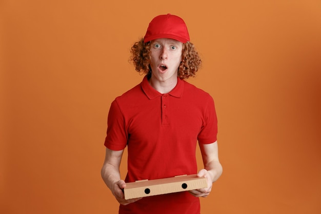 Pracownik dostawy w czerwonej czapce pustej koszulce mundurze trzymającym pudełko po pizzy patrząc na kamerę zdumiony i zaskoczony stojąc na pomarańczowym tle