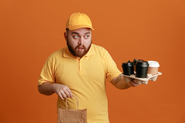 Pracownik Dostawy W Mundurze Pustej Koszulki żółtej Czapce, Trzymający Filiżanki Kawy I Papierową Torbę, Patrząc Na Bok Zdezorientowany, Stojąc Na Pomarańczowym Tle