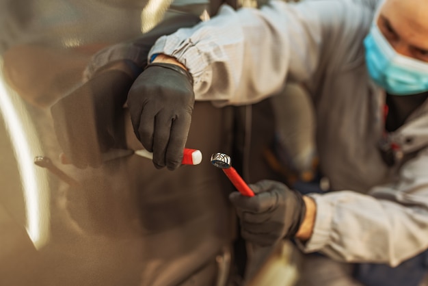 Pracownik fabryki samochodów w ochronnej masce medycznej naprawia małą wadę metalu za pomocą narzędzia ręcznego