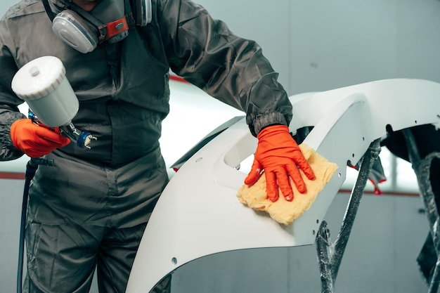 Pracownik mechanika samochodowego w odzieży ochronnej rozpylający białą farbę na część samochodu w warsztacie