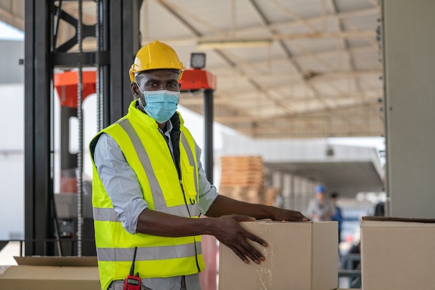 Pracownik mężczyzna nosi maskę na twarz w kamizelce odblaskowej i żółtym kasku, przygotowując wejście produktu do pudełka do wysyłki w fabryce magazynu
