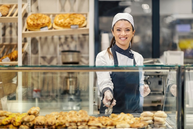 Pracownik piekarni sprzedaje ciasto i chleb w supermarkecie, uśmiechając się do kamery