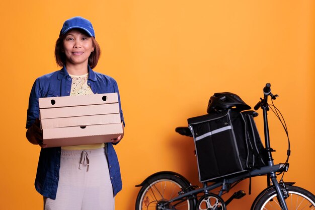 Pracownik restauracji ubrany w niebieski mundur trzymający stos pizzy dostarczającej klientowi na rowerze. Pracownik dostawy żywności stojący w studio z żółtym tłem. Usługi gastronomiczne i transport