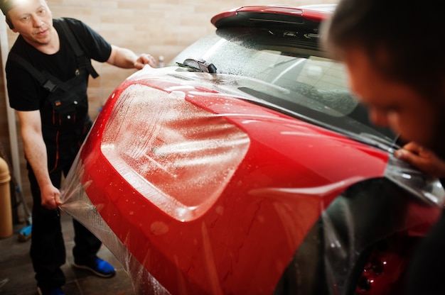 Pracownik serwisu samochodowego nałożył folię przeciw żwirowi na czerwoną karoserię samochodu w warsztacie samochodowym detailingowym