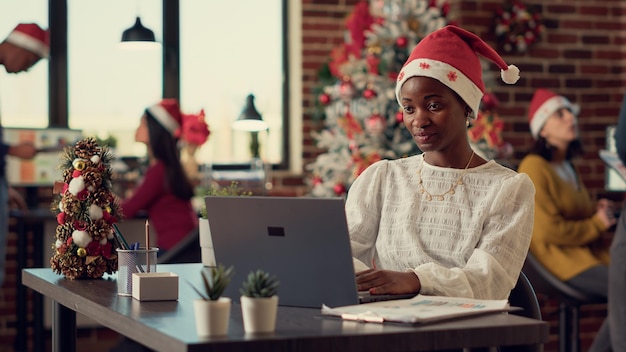 Pracownik startowy pracujący na laptopie podczas ferii zimowych, siedzący przy biurku ze świątecznymi dekoracjami. Kobieta w santa hat korzysta z komputera przed świętowaniem wigilii. Strzał ze statywu.