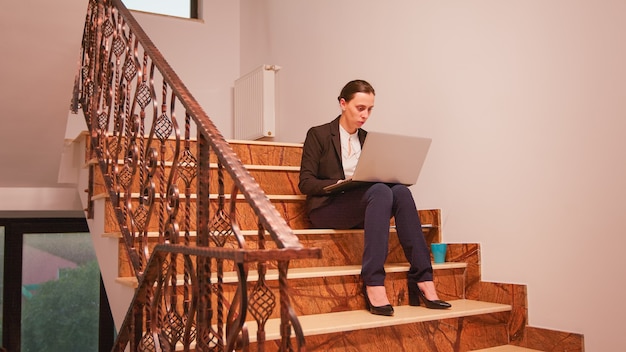 Pracownik Za Pomocą Laptopa Przepracowany W Terminie, Siedząc Na Schodach W Finansach Firmy, Podczas Gdy Koledzy Opuszczają Miejsce Pracy Biurowej. Profesjonalni Biznesmeni Pracujący W Nowoczesnym Budynku Finansowym