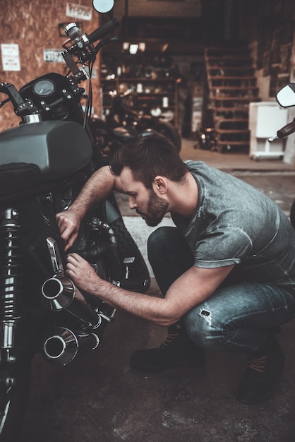 Pracuje w jego garażu. Pewny siebie młody człowiek naprawiający motocykl w pobliżu swojego garażu