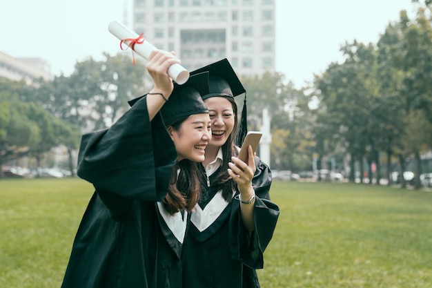 Prawdziwy styl życia w dniu ukończenia studiów. dwie szczęśliwe kobiety studentów przyjaciele w końcu absolwent w czapce suknie trzymać dyplom rozmawiając przez telefon głośnomówiący krzyczeć świętując wesoły. dziewczyny bawią się telefonem komórkowym