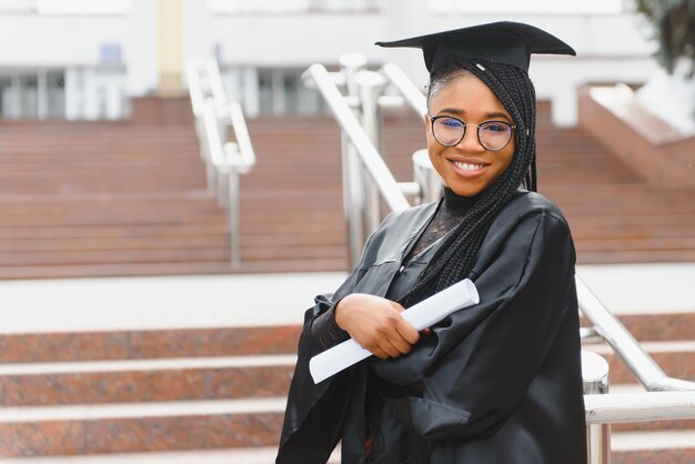 Pretty African studentka w czapce i sukni ukończenia szkoły przed budynkiem szkoły