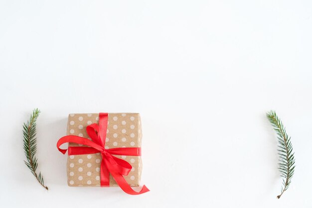 Prezent na Boże Narodzenie i nowy rok owinięty w kropkowany papier rzemieślniczy i związany czerwoną wstążką na białym tle ręcznie robiony prezent i wystrój gałęzi jodłowych
