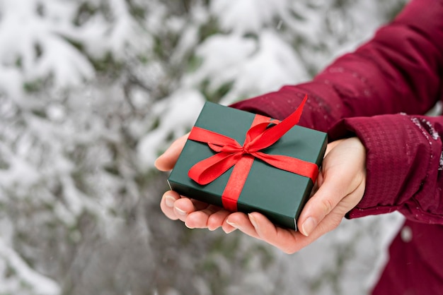 Prezent w zielonym pudełku z czerwoną wstążką w kobiecych rękach na ośnieżonym drzewie w zimowym tle bożego narodzenia