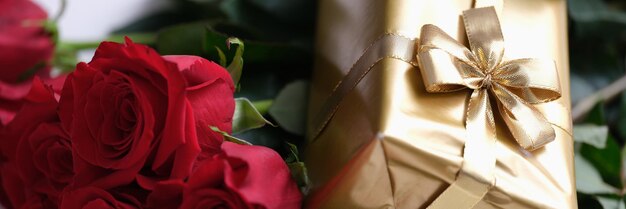 Prezent w złotym opakowaniu z kokardą leżącą na zbliżeniu bukietu czerwonych róż