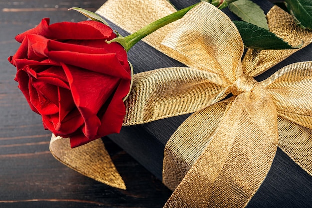 Prezent zapakowany w czarny papier i złotą wstążkę. na górze pudełka znajduje się luksusowa czerwona róża. Koncepcja drogiego prezentu na świąteczne Walentynki lub Dzień Kobiet 8 marca