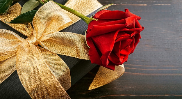 Prezent zapakowany w czarny papier i złotą wstążkę. na górze pudełka znajduje się luksusowa czerwona róża.