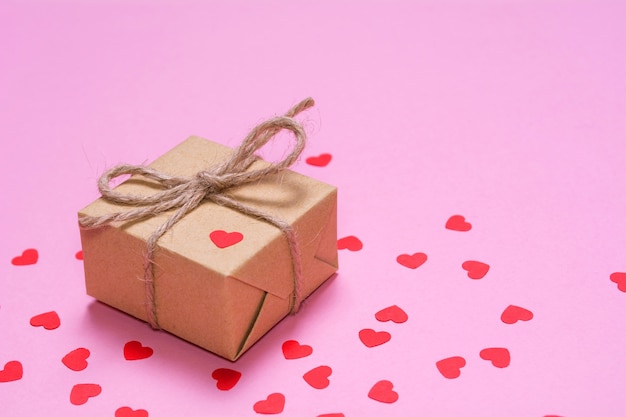 Prezent zawinięty w papier pakowy na różowym tle. Papierowe czerwone serca na pudełku prezentowym.