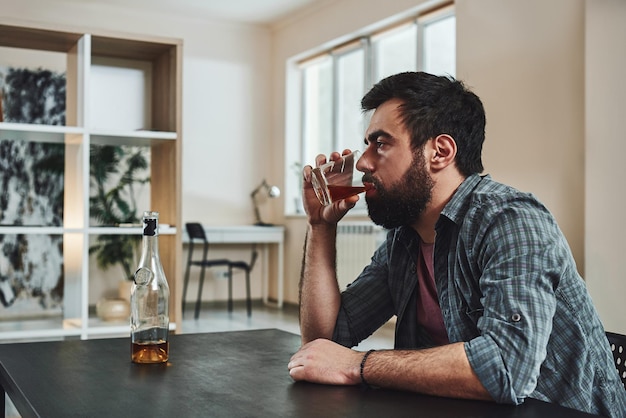 Problemy z piciem. Przygnębiony brodaty mężczyzna siedzi przy stole ze szklanką whisky w ręku. Myśli o problemach w pracy i kłopotach w związkach podczas picia alkoholu. Widok z boku