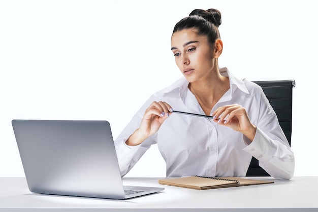 Proces biznesowy i koncepcja pracy z ładną młodą kobietą siedzącą przy białym biurku, patrzącą na ekran laptopa i trzymającą ołówek w rękach na abstrakcyjnym tle białej ściany