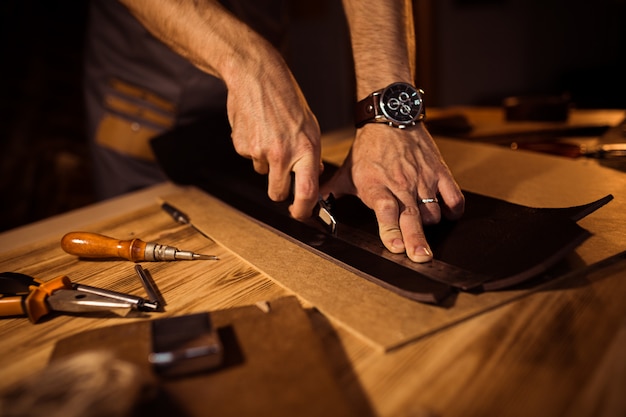 Proces pracy skórzanego paska w warsztacie skórzanym. Mężczyzna trzyma wykonywać ręcznie narzędzie i działanie. Garbarz w starej garbarni. Drewniany stół tło.