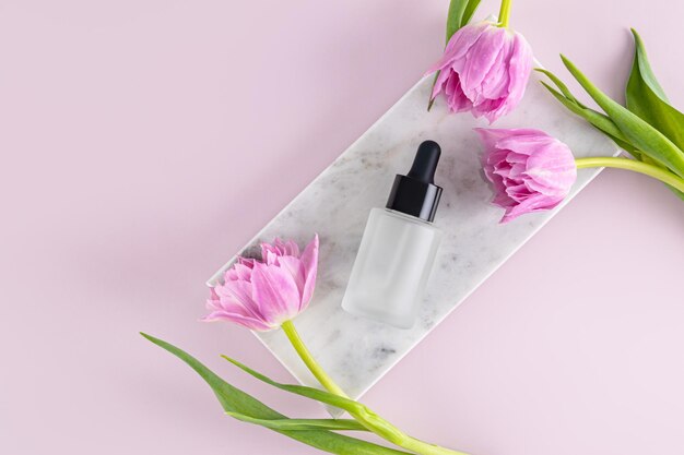 Produkt kosmetyczny serum olejkowe do pielęgnacji skóry twarzy i ciała w matowym butelce z kroplówką leży na marmurowym podium z delikatnymi tulipanami