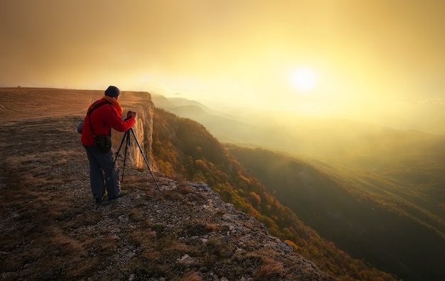 Profesjonalista w górach. Fotograf przyrody robi zdjęcia lustrzanym aparatem na szczycie skały. Marzycielski mglisty krajobraz, wiosenny pomarańczowy mglisty wschód słońca w pięknej dolinie poniżej.