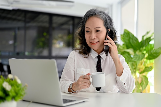 Profesjonalna azjatycka bizneswoman pracująca w biurze rozmawiająca przez telefon i popijająca kawę