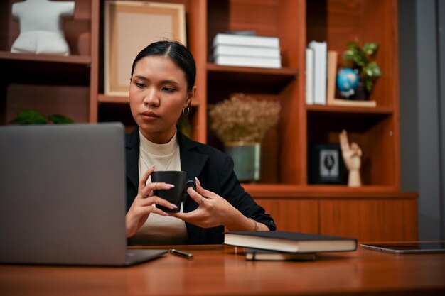 Profesjonalna azjatycka bizneswoman trzymająca filiżankę kawy i jednocześnie patrząc na ekran laptopa