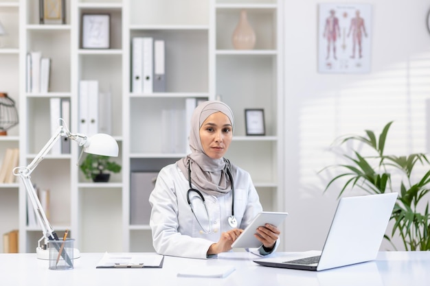 Profesjonalna muzułmańska lekarzka w hidżabie i białym płaszczu pracująca w swoim biurze za pomocą tabletu